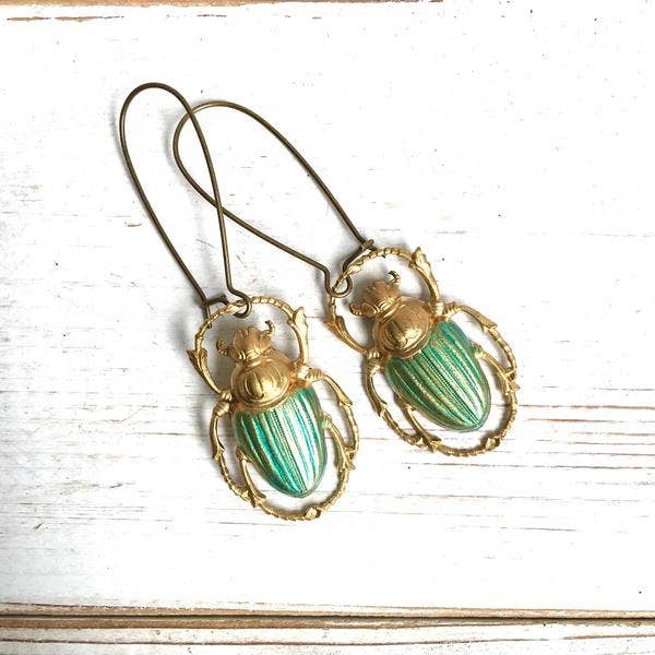 The Green Beetle Earrings are vintage beetle-shaped earrings with gold outlines and green striped detailing on their backs, beautifully displayed on a white wooden surface.