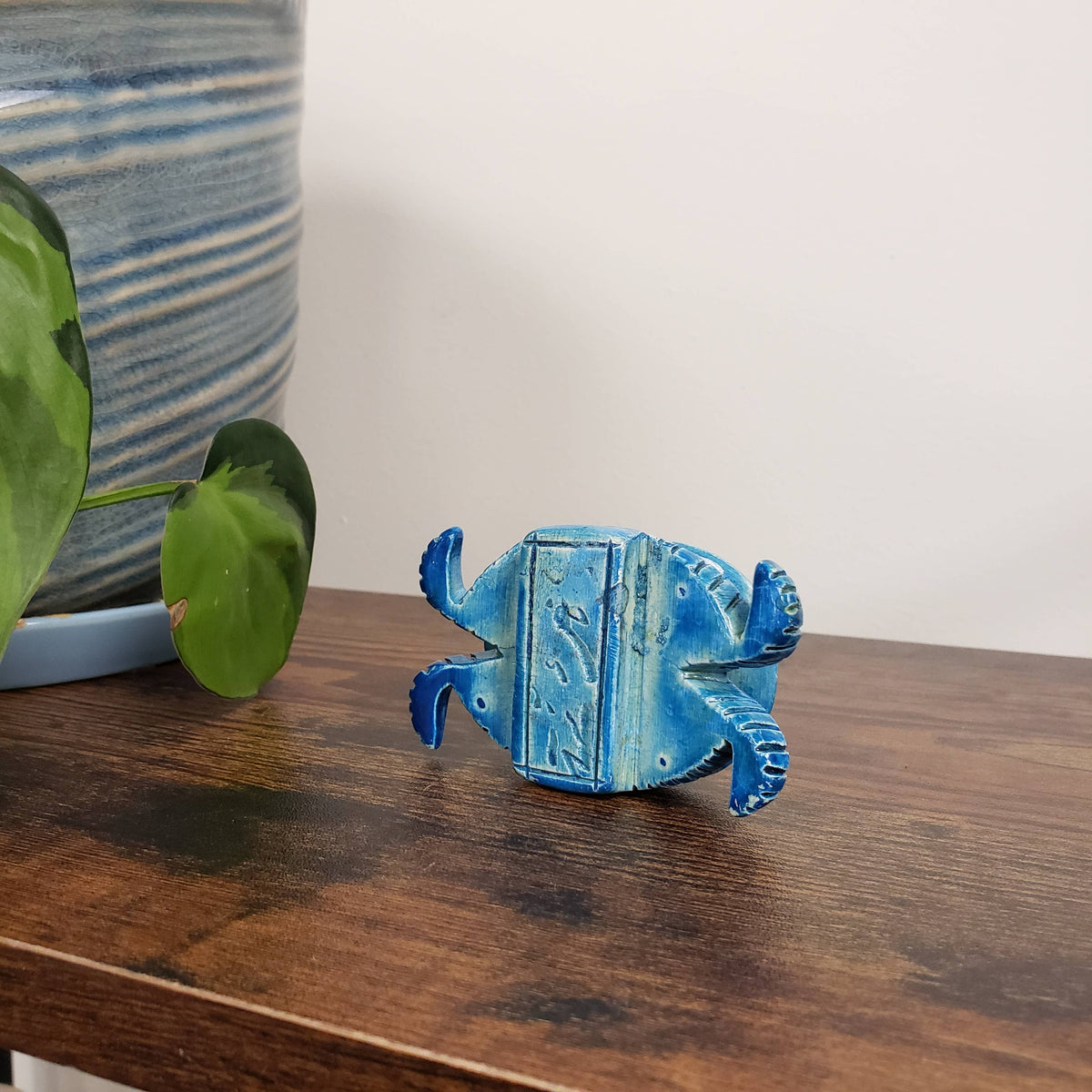 A blue ceramic crab figurine with decorative markings sits on a wooden shelf beside a green plant, a striped blue vase, and a Soapstone Turtle Statue in blue (1.25″, made in Egypt) shaped like an ancient amulet.