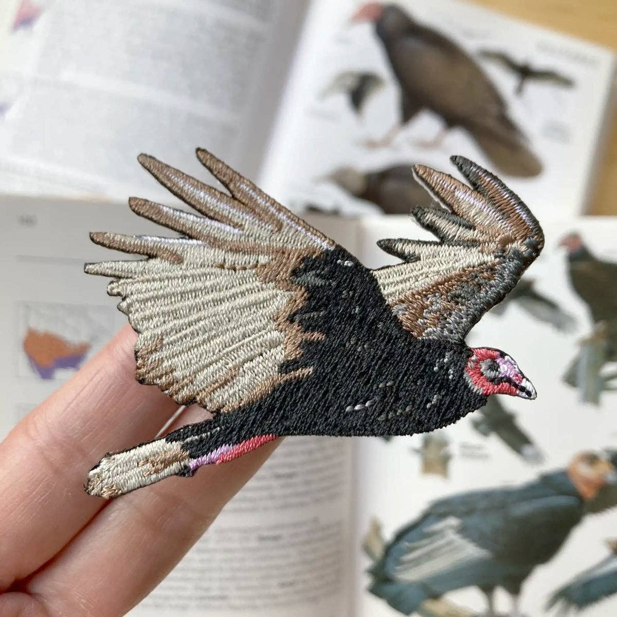 A hand holds the Turkey Vulture Patch, embroidered with brown, black, and cream feathers and a red head—ideal for scavenger fans. In the background, an open bird guidebook displays vulture images.