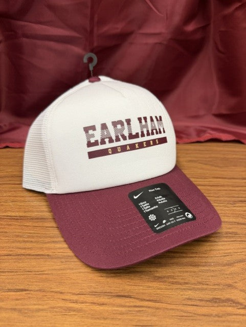 A Nike Foam Trucker Hat in white and maroon, featuring "EARLHAM QUAKERS" on the foam front, sits on a wooden surface with a maroon fabric background.