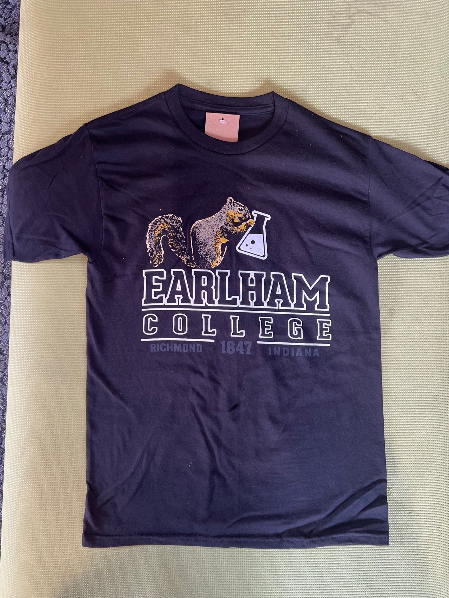 The Squirrel Lab Tee features a black squirrel holding a beaker with "EARLHAM COLLEGE, RICHMOND 1847 INDIANA" text. The 23/24 edition is shown laid flat on a light green background.