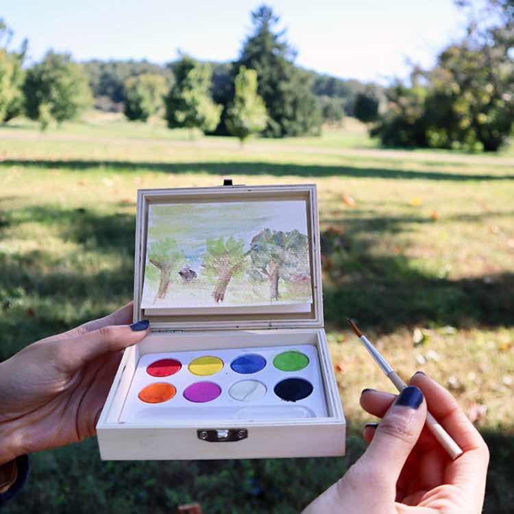 A person uses the Huckleberry Landscape Paint Kit and brush outdoors on grass, painting trees on a watercolor paper pad, with the artwork matching the landscape in the background.
