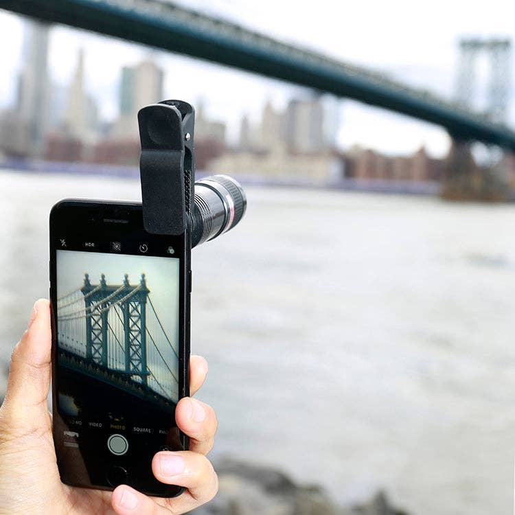 A hand holds a smartphone with the Phone Telescope clipped on, capturing a sharp photo of a suspension bridge over a river and the blurred city skyline beyond, all magnified 8x for detailed clarity.