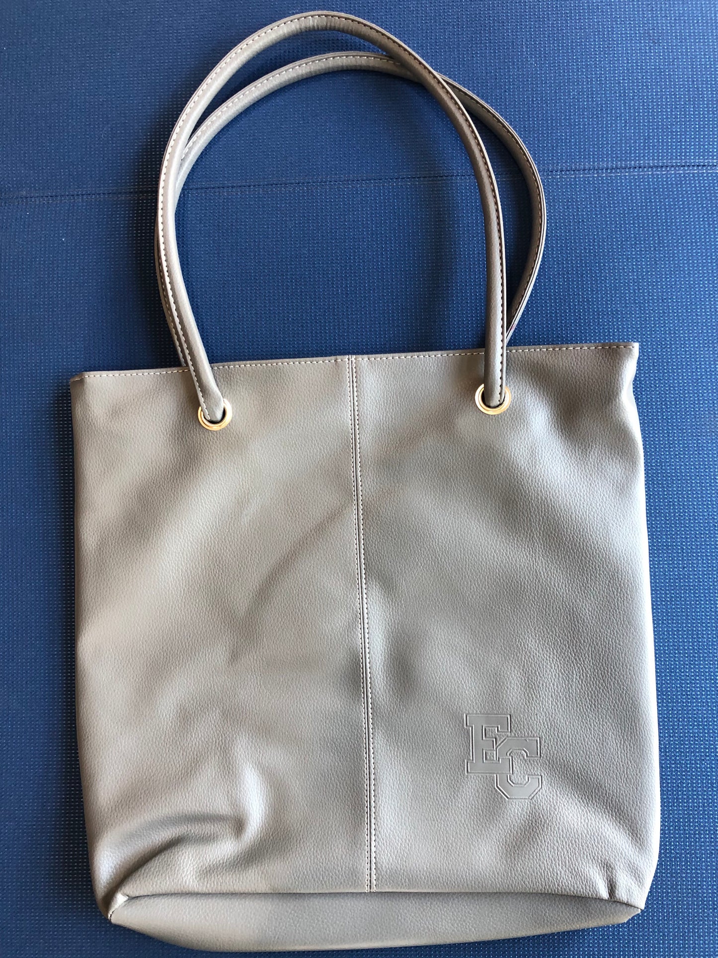 The EC Debossed Tote Bag is a light gray leather tote with two handles, gold grommets, and an embossed EC monogram at the lower right corner, shown on a blue textured surface.