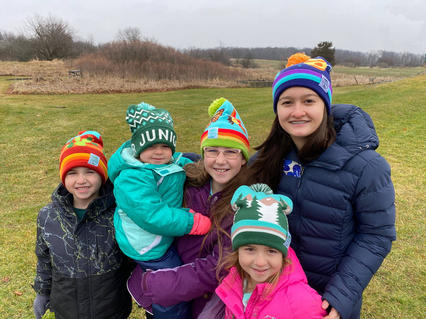 Five smiling children wearing colorful winter hats and jackets—including a solar system hat and the Junior Ranger Gear Night Sky Stocking Cap—stand together outdoors on a grassy field with cloudy skies and barren trees in the background.
