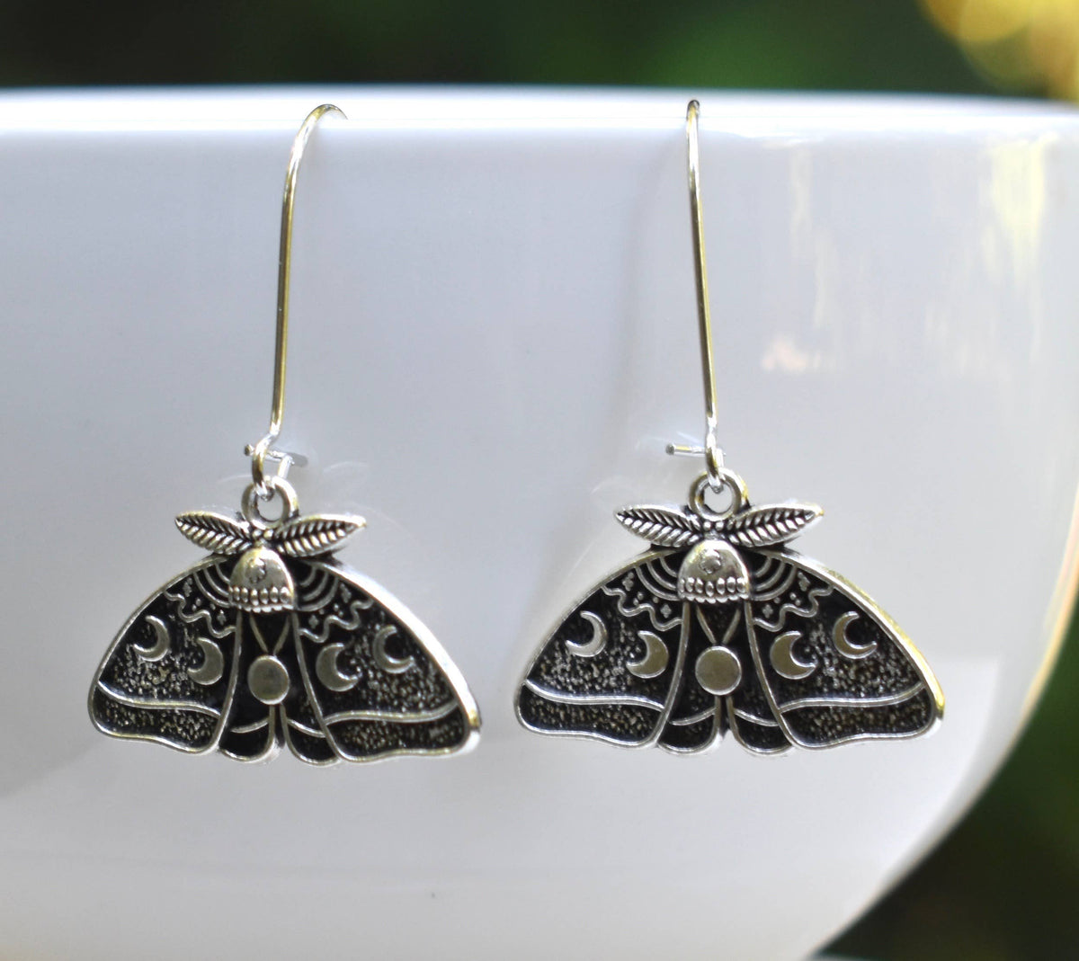 The Luna Moth Earrings feature intricate silver designs and dangle from hooks, beautifully displayed on the edge of a white bowl with a softly blurred green background.
