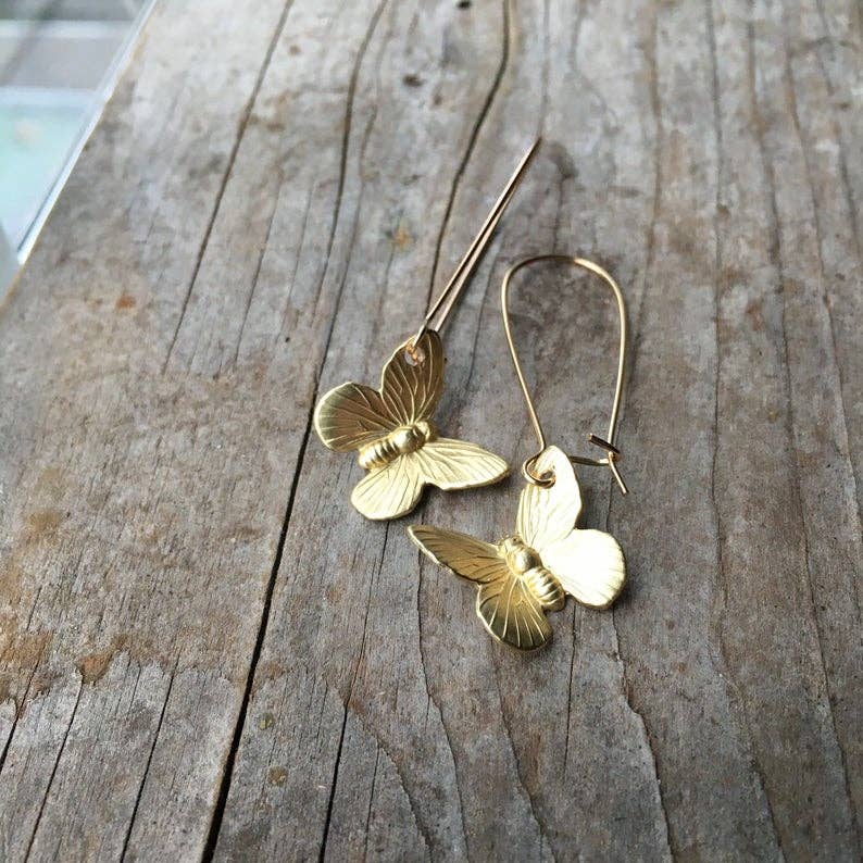 A pair of SmallButterfly Earrings with delicate gold butterfly charms rests on a weathered wooden surface, capturing the essence of nature-inspired jewelry.