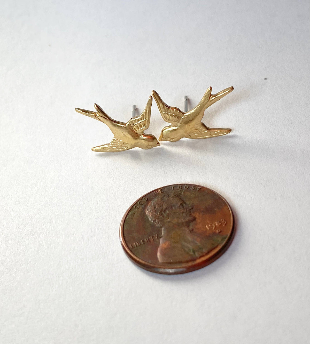 Two Flying Sparrow Earrings in a gold bird-shaped stud design are placed above a US dime on a white surface, showcasing their size. These golden brass Flying Sparrow Earrings make lovely nature-inspired jewelry, perfect for a woodland wedding or everyday wear.