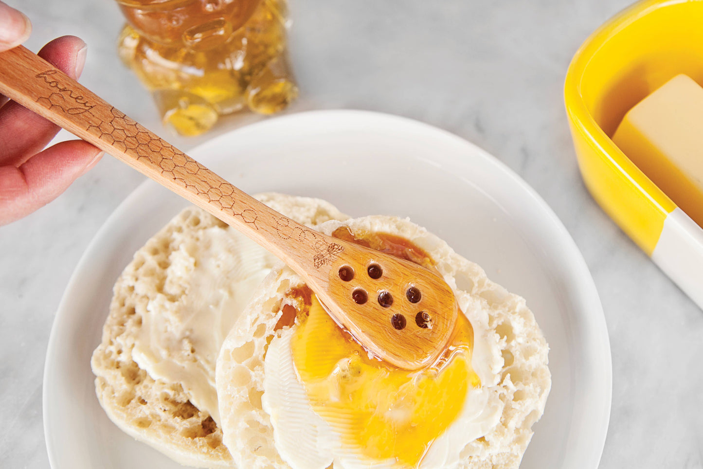 A hand uses the TALISMAN DESIGNS Honey Bee Honey Spreader and Dipper to drizzle honey onto a buttered English muffin on a white plate, with a bear-shaped honey jar and yellow butter dish in the background.