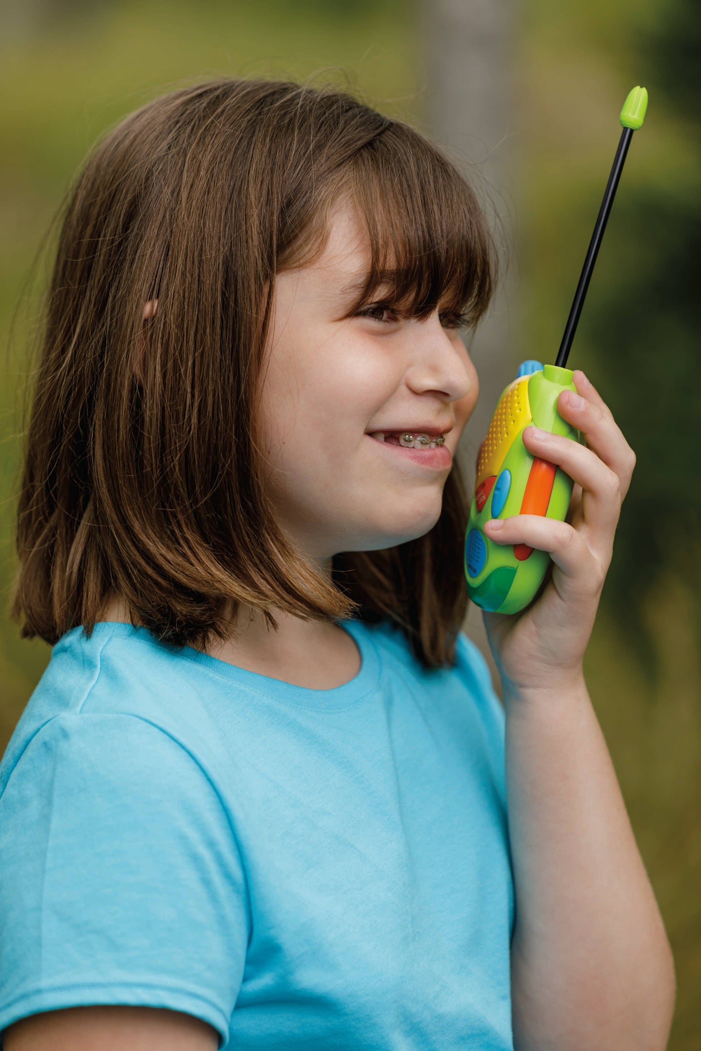 A smiling young girl with brown hair and braces, wearing a light blue shirt, uses the Outdoor Discovery Walkie Talkies with volume control outdoors.