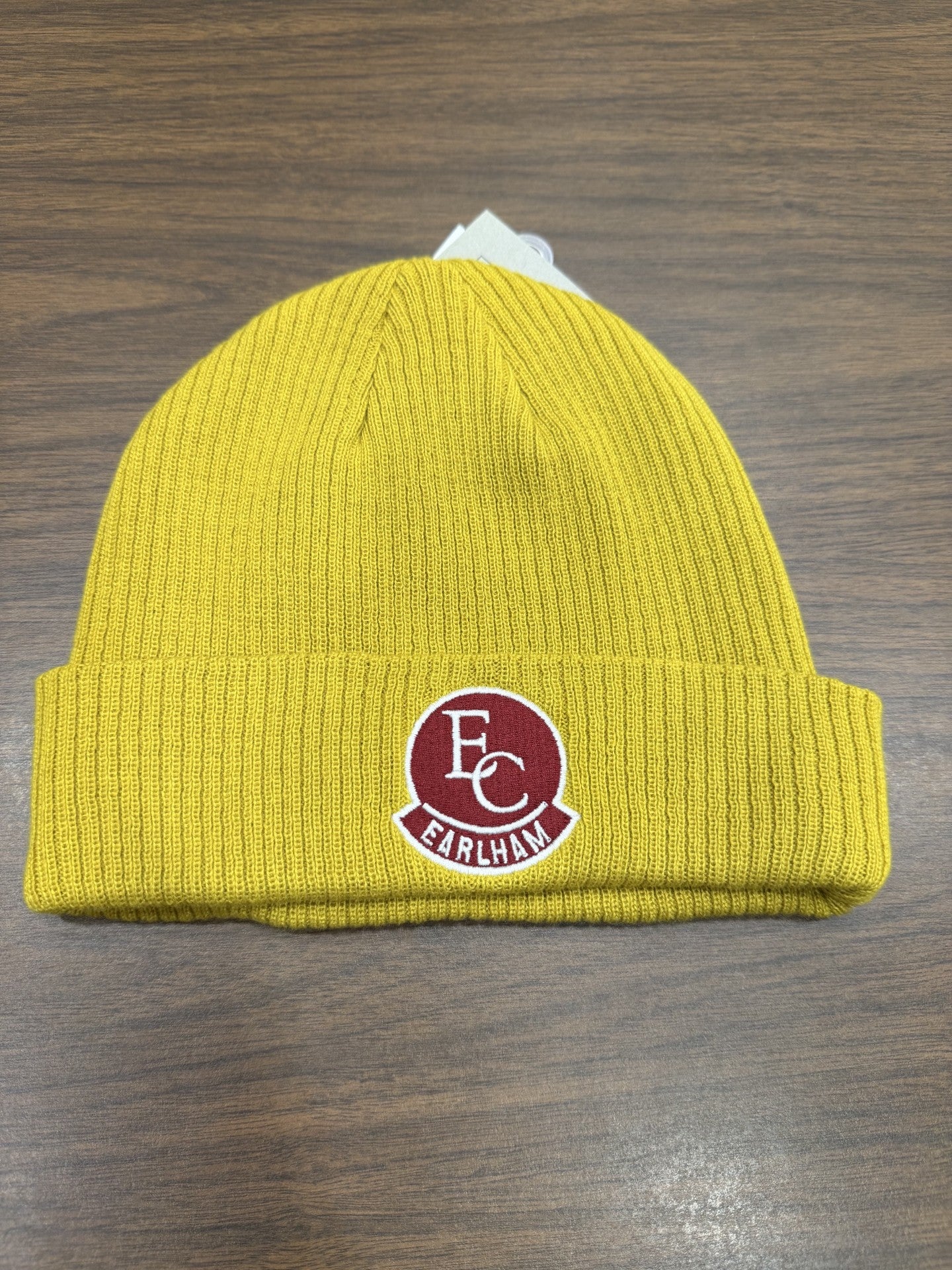 The Golden Rollup Beanie, a yellow knit hat with a folded brim and a red and white EC EARLHAM patch on the front, is placed on a wooden surface.