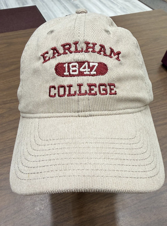 A beige “for good.” Corduroy Cap with EARLHAM 1847 COLLEGE embroidered in red on the front, displayed on a wooden table indoors.