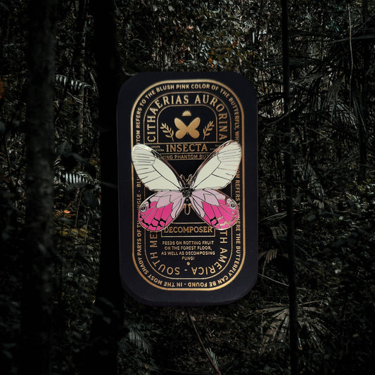 A can with a butterfly illustration and ornate label stands upright in a dark, dense forest with tall trees and lush foliage in the background, evoking the delicate allure of the Blushing Phantom Butterfly Educational Insect Enamel Pin—a rare collectible accessory perfect for discerning enthusiasts.