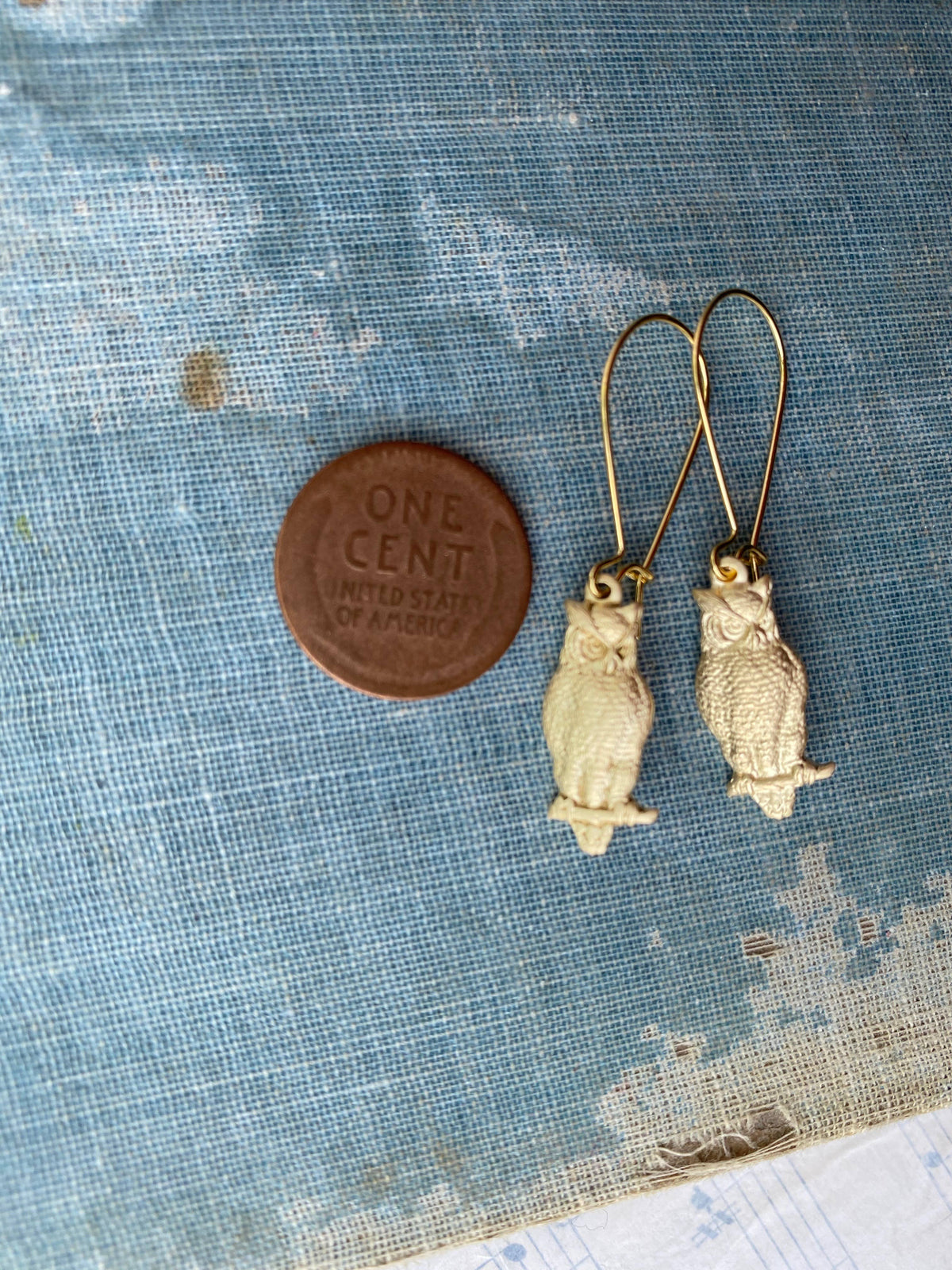 The Wise Owl Earrings, a charming small vintage pair with gold hooks, are displayed beside a one cent coin on a textured blue surface.