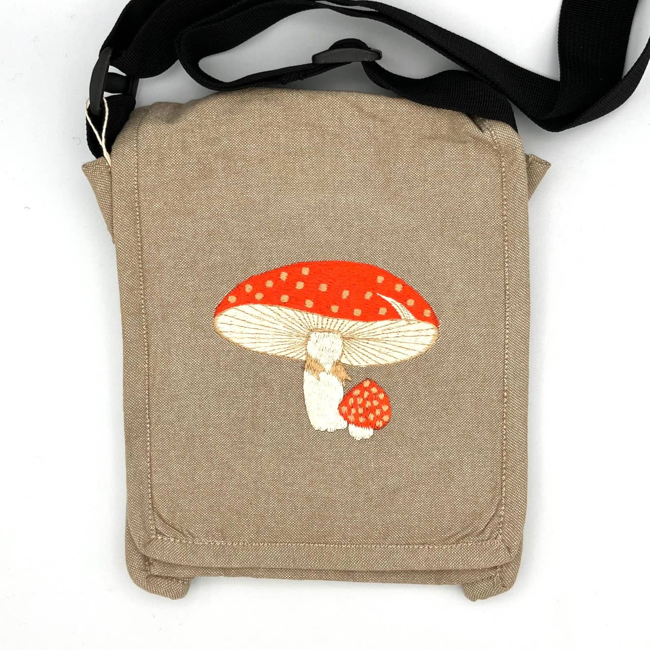 The Fly Agaric Mushroom Field Bag is a small handmade cotton shoulder bag in tan with a black strap. Its front flap displays embroidered red and white spotted mushrooms, making it a charming companion for birdwatching outdoors.