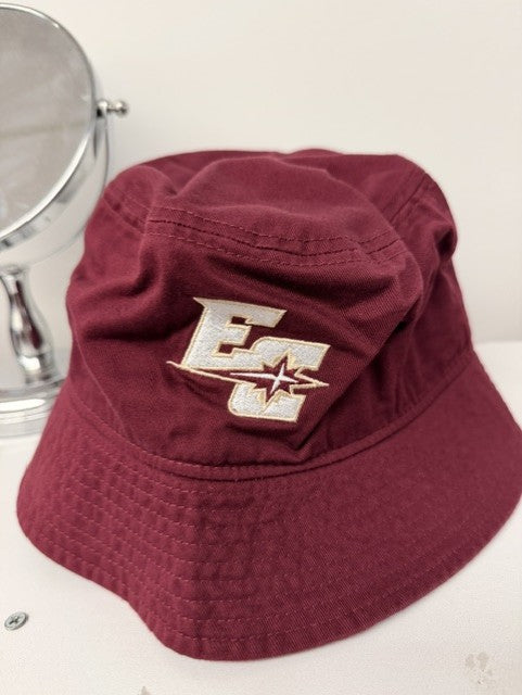 A Nike Bucket Hat in maroon, featuring an embroidered EC logo with a star, is placed next to a round mirror on a white surface.