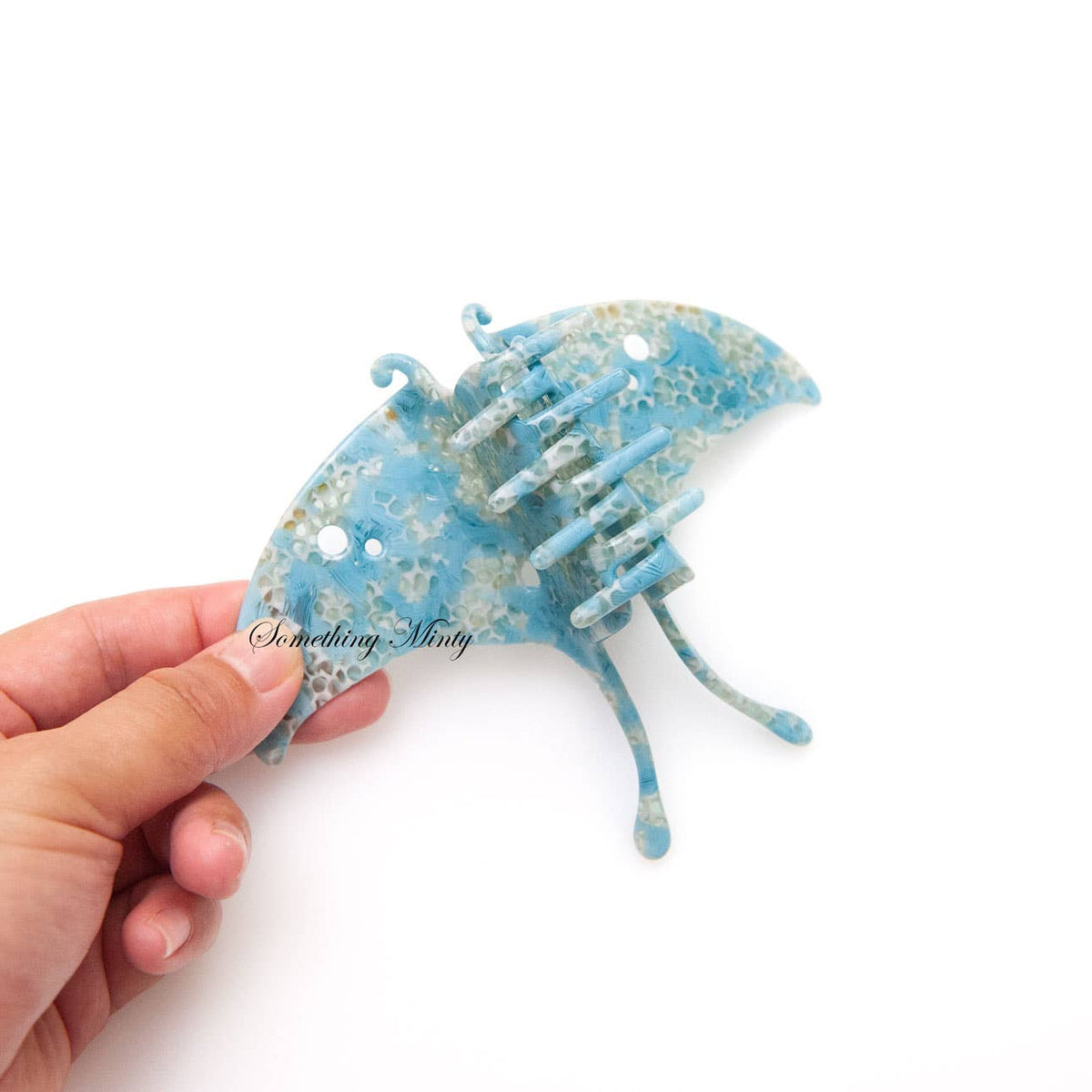 A hand holding a blue, moth-shaped Luna Moth Hair Claw Clip made of acetate with a marbled pattern.
