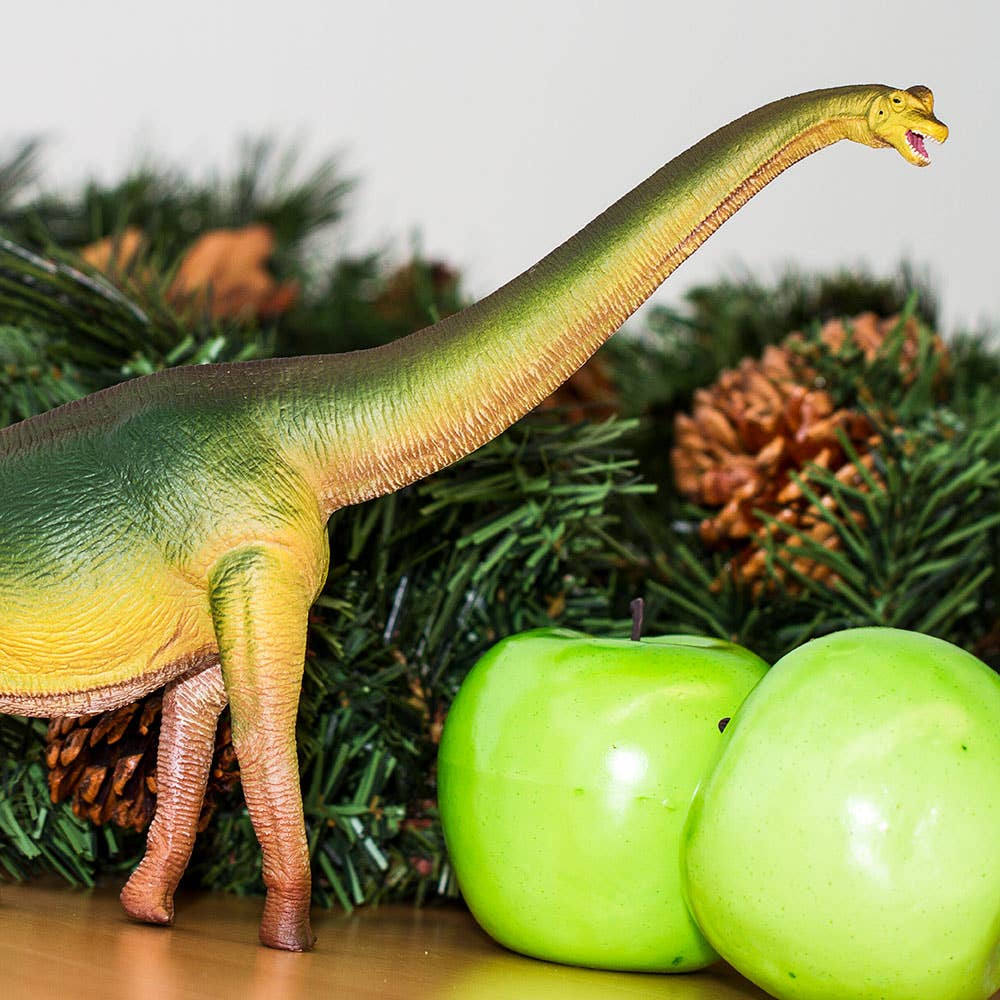 A Brachiosaurus stands beside two green apples, with pine branches and pinecones in the background.