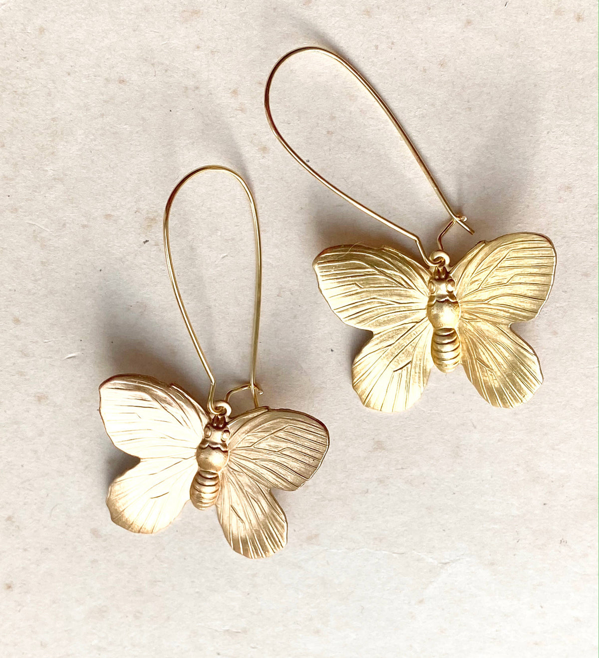 Two Butterfly Earrings, featuring gold butterfly-shaped designs with detailed wings and long, thin hooks, are displayed on a light beige surface—elegant statement pieces perfect for any occasion.