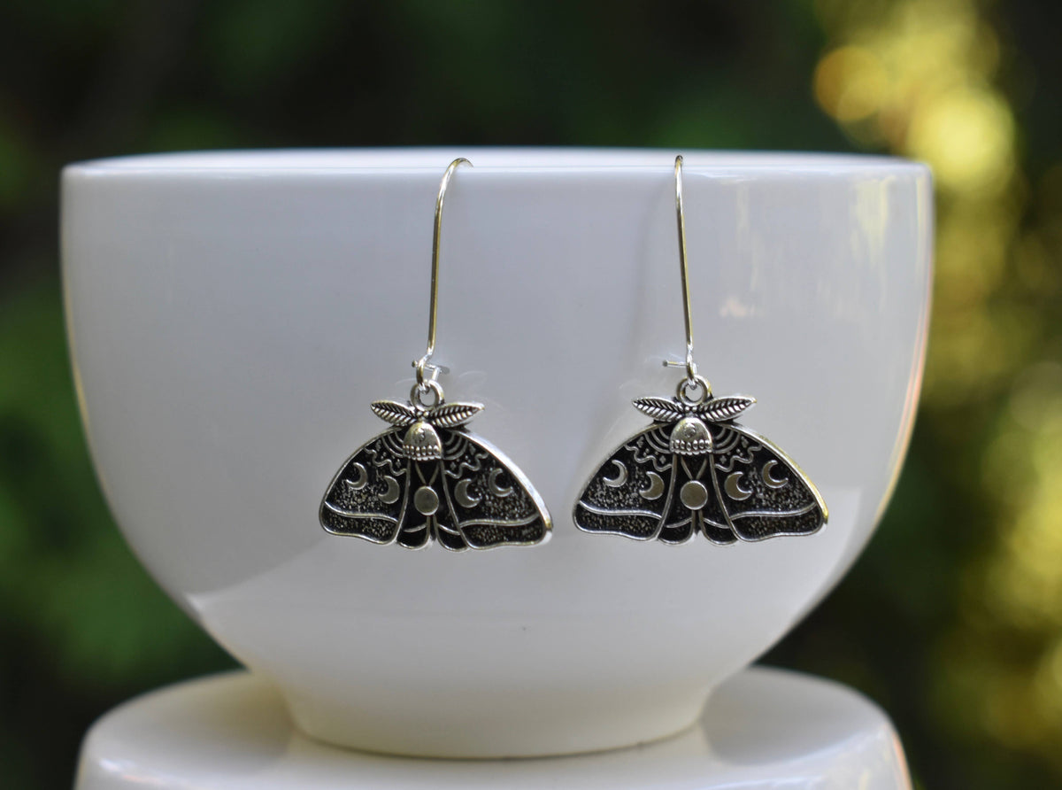 The Luna Moth Earrings, crafted in silver with intricate detailing, hang elegantly from a white bowl against a softly blurred green background.