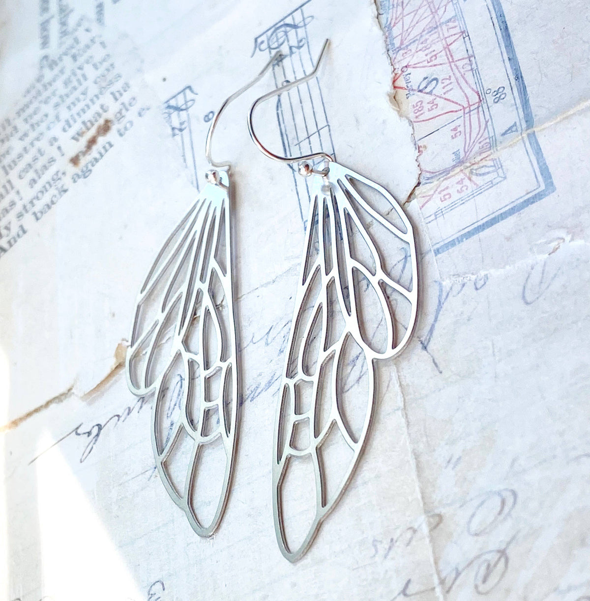 Silver Lace Cicada Wings earrings, artfully crafted in silver with delicate lace detailing, rest atop vintage paper featuring handwritten notes and faded blueprints in the background.
