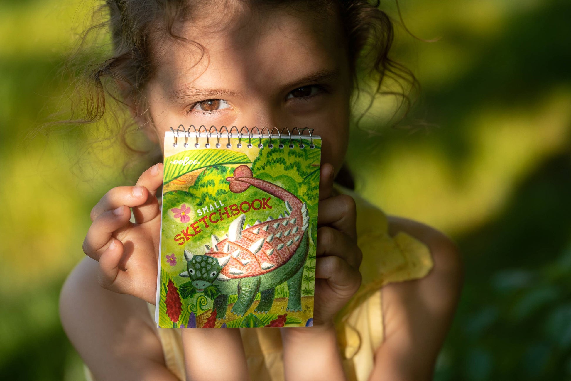 A young child in a yellow dress holds up a Small Sketchbooks Dino Assortment, featuring a colorful dinosaur illustration and 'SKETCHBOOK' on the cover, as sunlight casts shadows across their face and background.