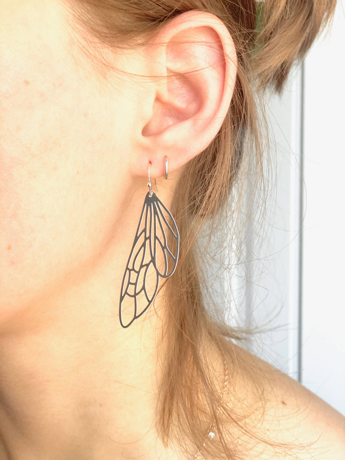A close-up of a person’s ear wearing a large, delicate Silver Lace Cicada Wings earring shaped like an insect wing, with light brown hair partially covering the ear.