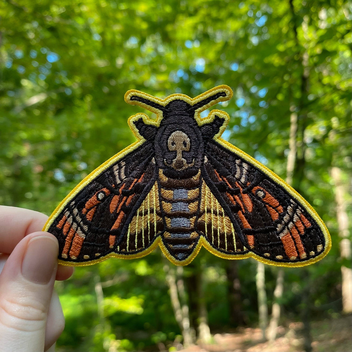 A Death’s-Head Hawkmoth Iron-On Embroidered Patch featuring a skull pattern on its back, held against a blurred green forest background.