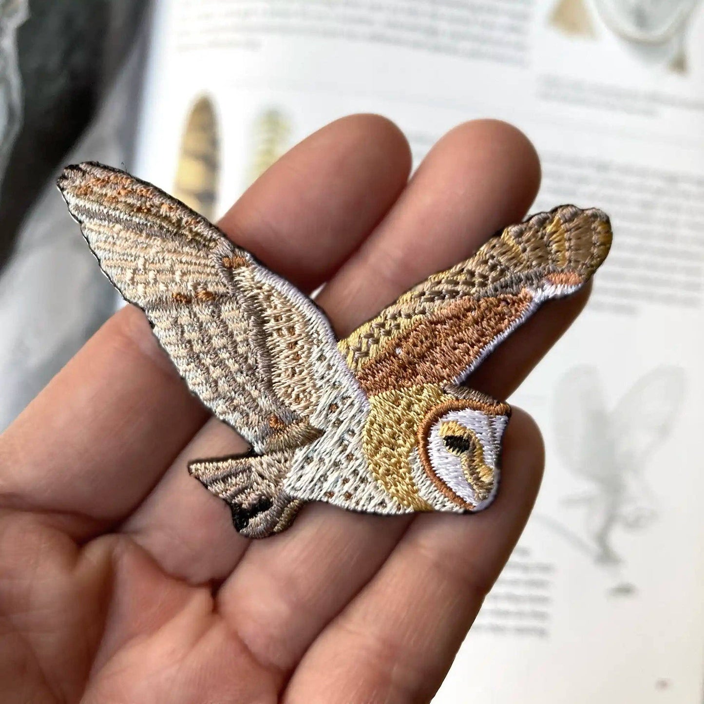 A hand holds the Barn Owl Patch, an embroidered patch shaped like a flying owl with outstretched wings in brown, cream, and white. A blurred open book with illustrations is seen in the background.