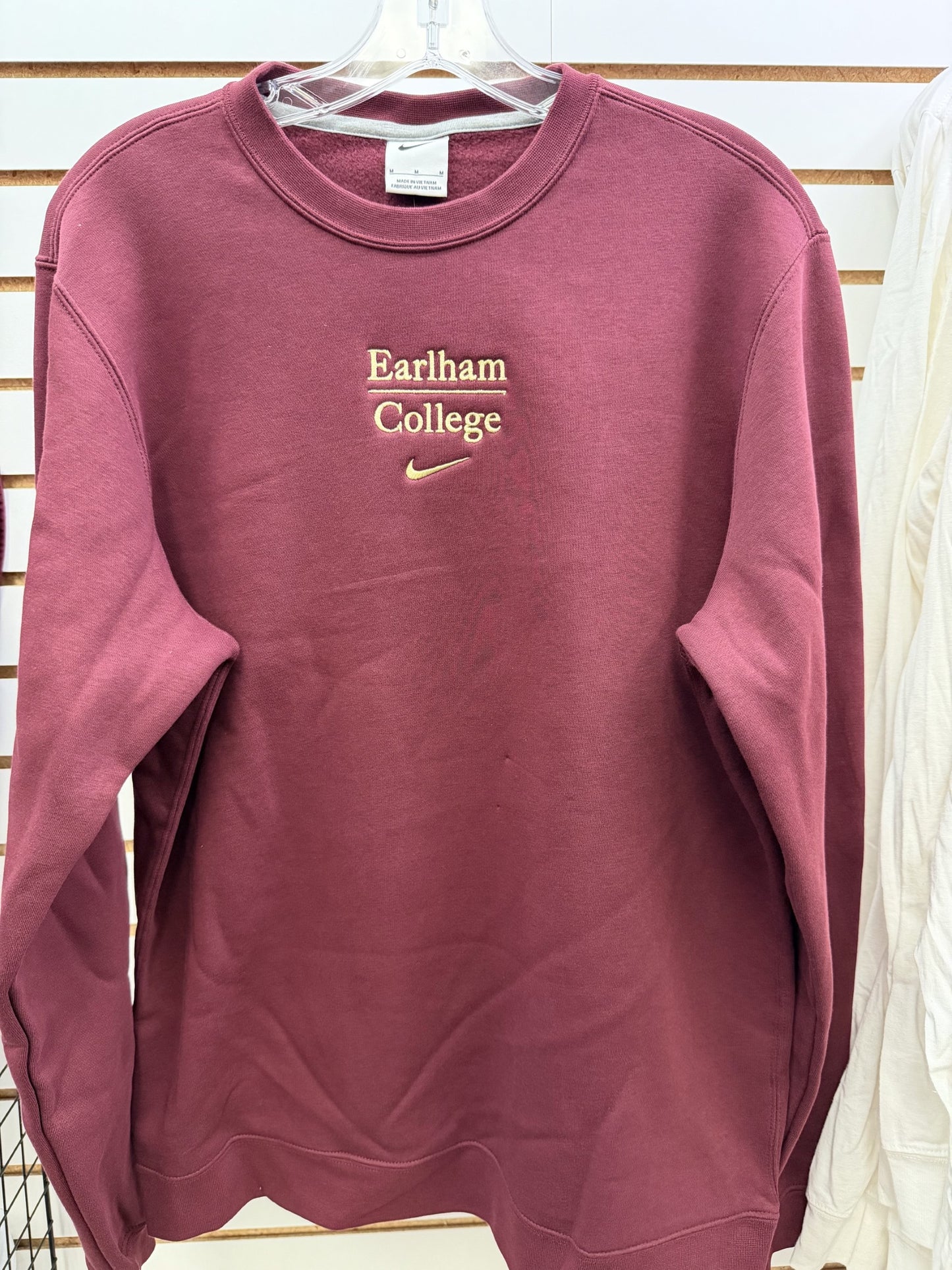 A Nike Embroidered Club Fleece sweatshirt in maroon, featuring "Earlham College" stitched in cream lettering, hangs on display against a white slat wall.
