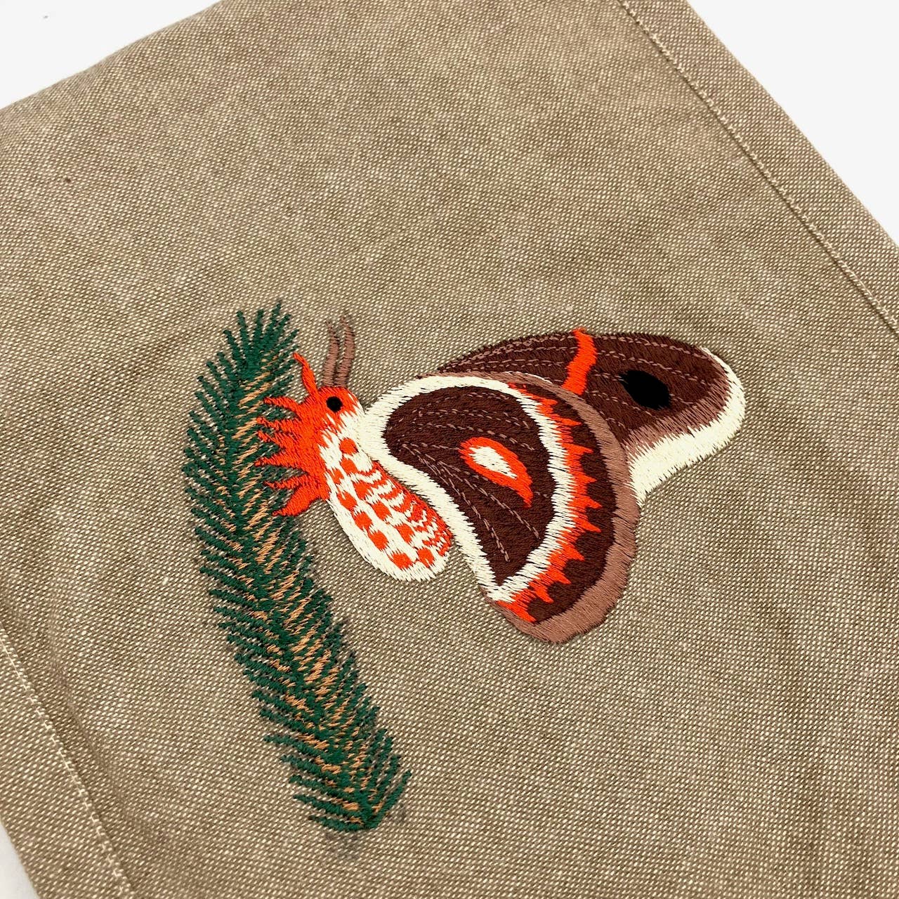 The Cecropia Moth Field Bag features a handmade, fair trade embroidered design of a brown, red, and white moth beside green pine needles on tan cotton—perfect for nature lovers seeking a unique accessory.