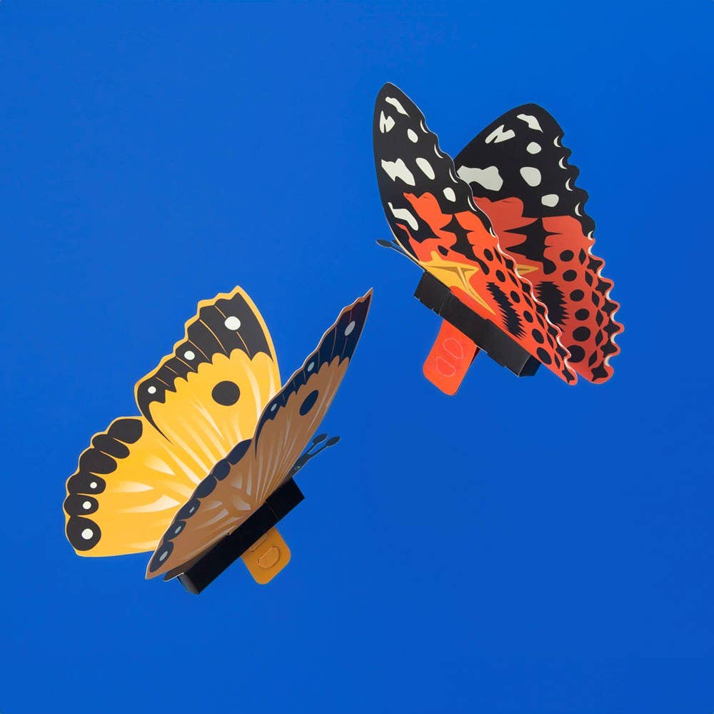 Two vibrant paper butterflies, made with the Create Your Own Fluttering Butterflies kit, one yellow with black accents and one red with black and white spots, are displayed on a bright blue background.
