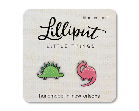 Dinosaur Earrings featuring one green stegosaurus and one pink brontosaurus stud, displayed on a card that reads “Lilliput Little Things, handmade in New Orleans.” These playful, hypoallergenic earrings are crafted with titanium posts for comfortable all-day wear.