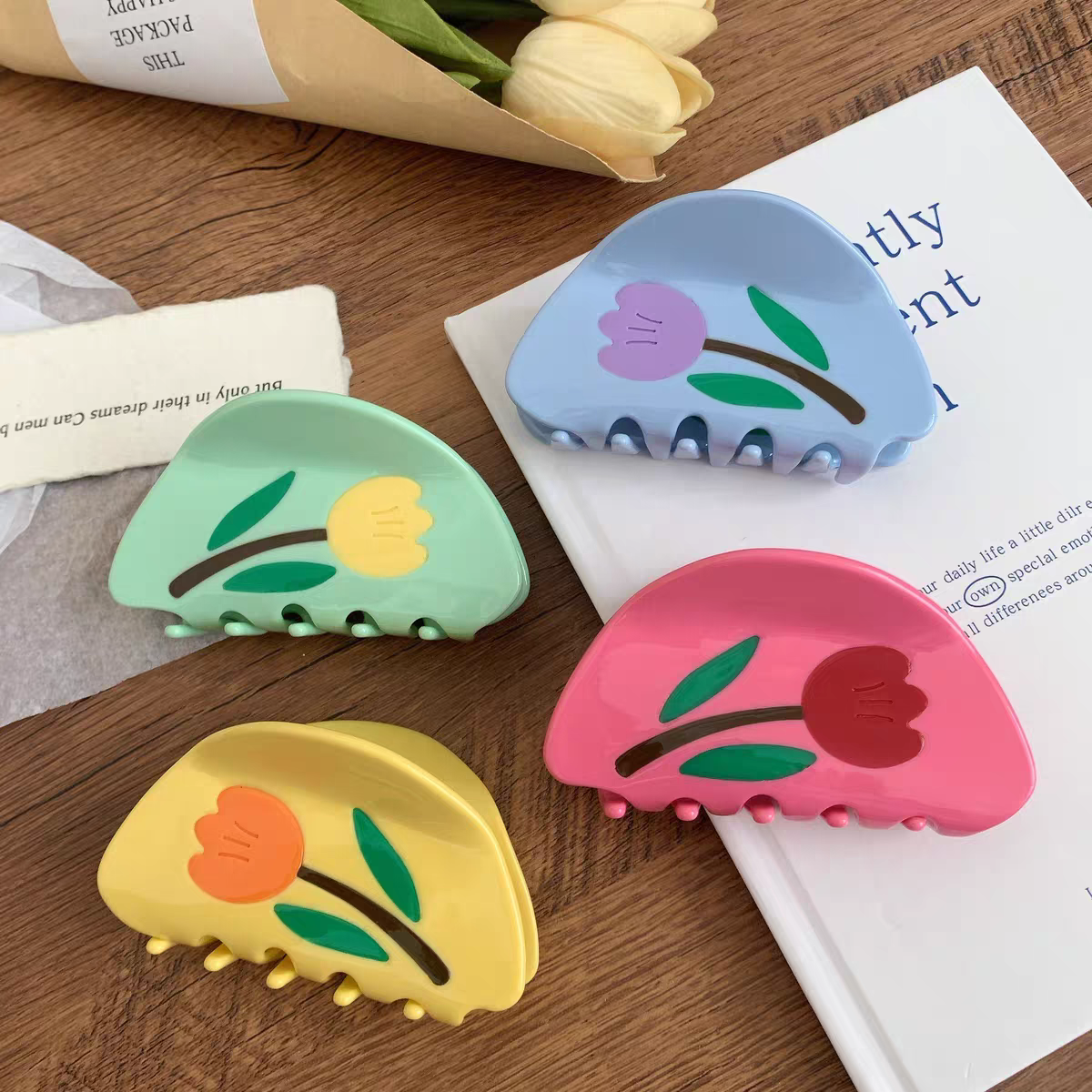 Four half-moon Tulip Hair Claw Clips in blue, green, yellow, and pink, each decorated with a single floral illustration.