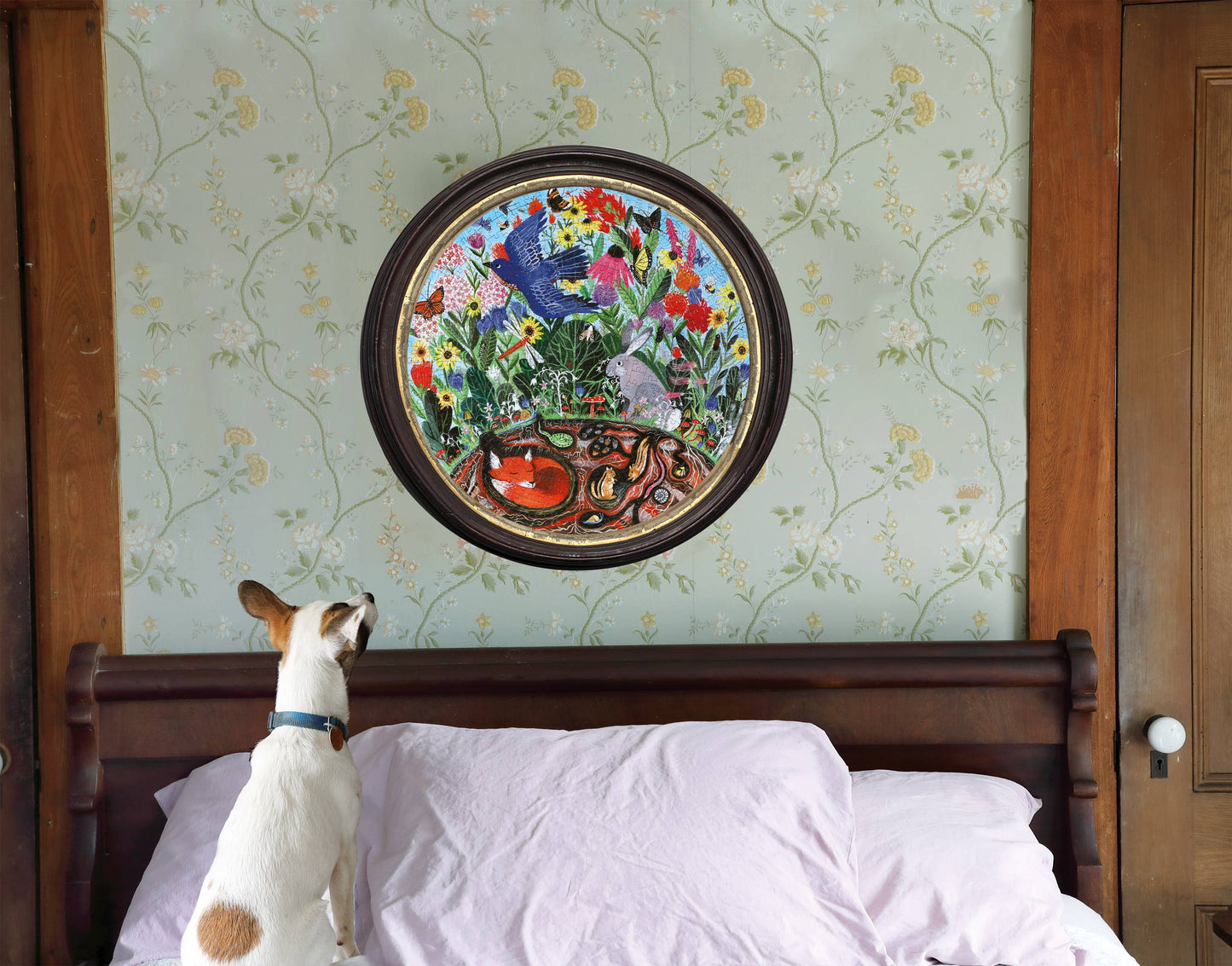 A small dog stands on a bed with rumpled white sheets, gazing up at the Rewilding 500 Piece Puzzle—a round, colorful illustration of garden animals, flowers, birds, and snakes—hanging on the wallpapered wall above.