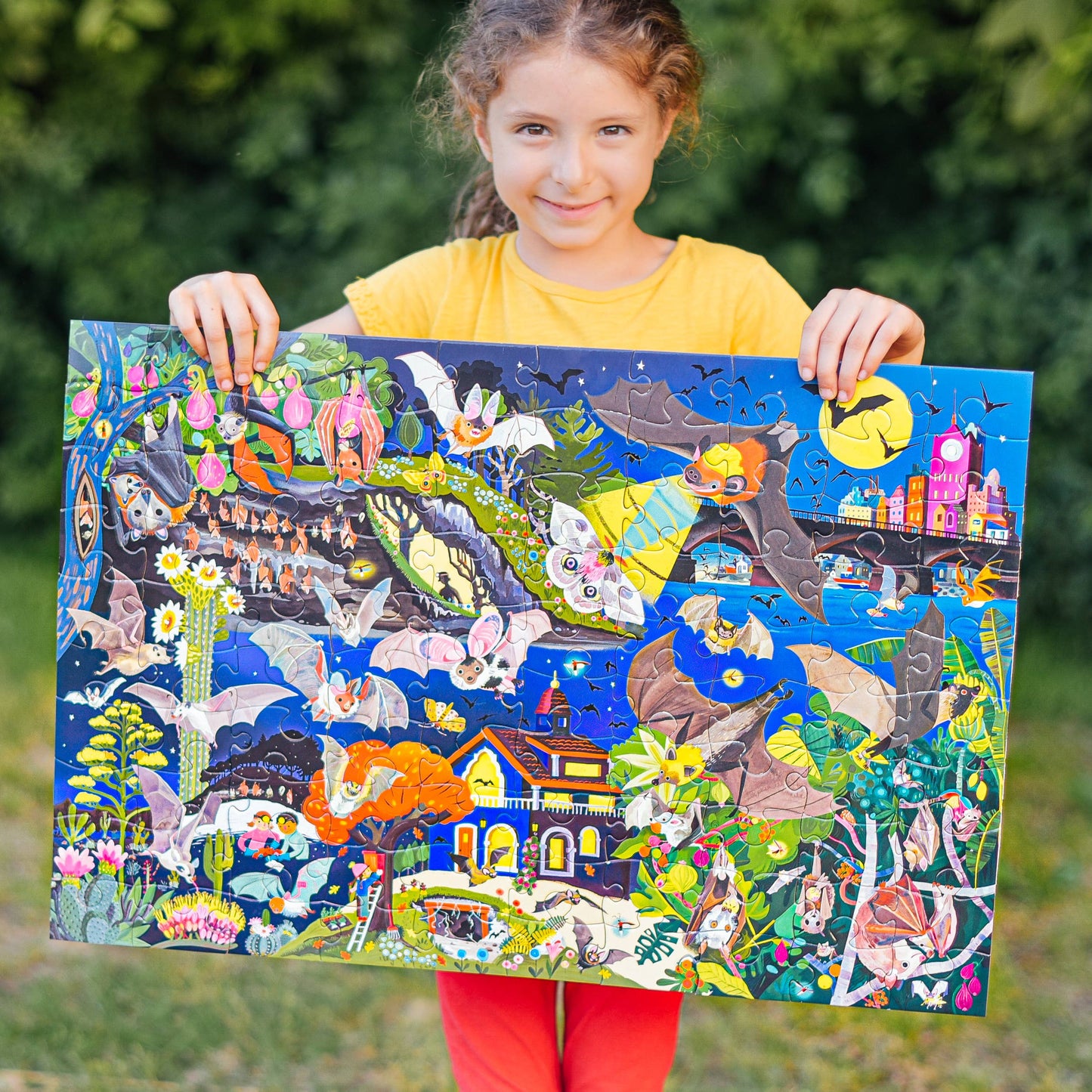 A smiling child in a yellow shirt and red pants holds up the "Love of Bats 100 Piece Glow in the Dark Puzzle," featuring vibrant animals, flowers, and colorful nature scenes, with blurred green foliage in the background.