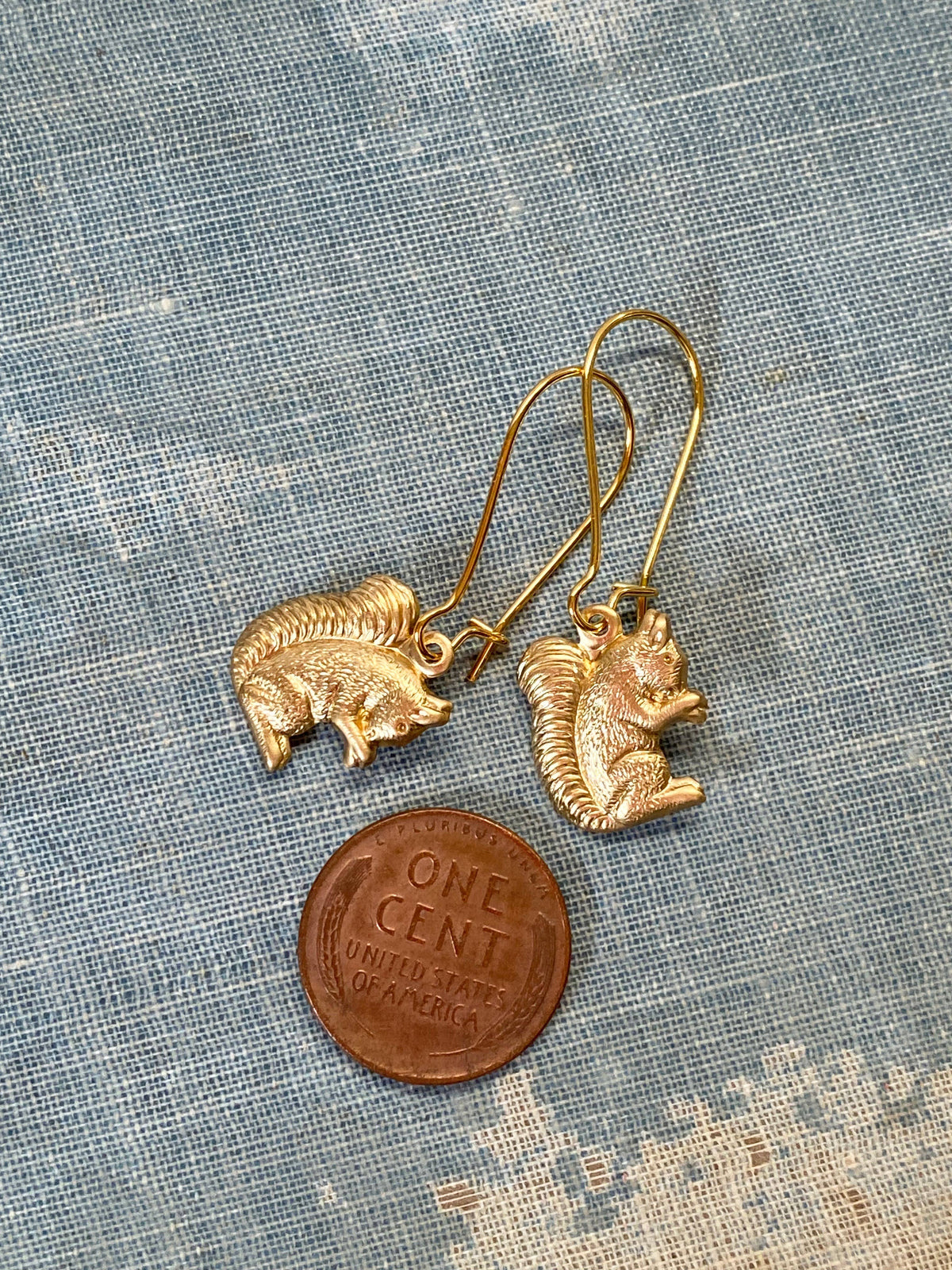 A pair of Woodland Forest Squirrel Earrings, shaped like playful gold squirrels, are displayed next to a U.S. one-cent coin on a blue patterned fabric background—perfect for anyone who loves whimsical woodland-themed jewelry.