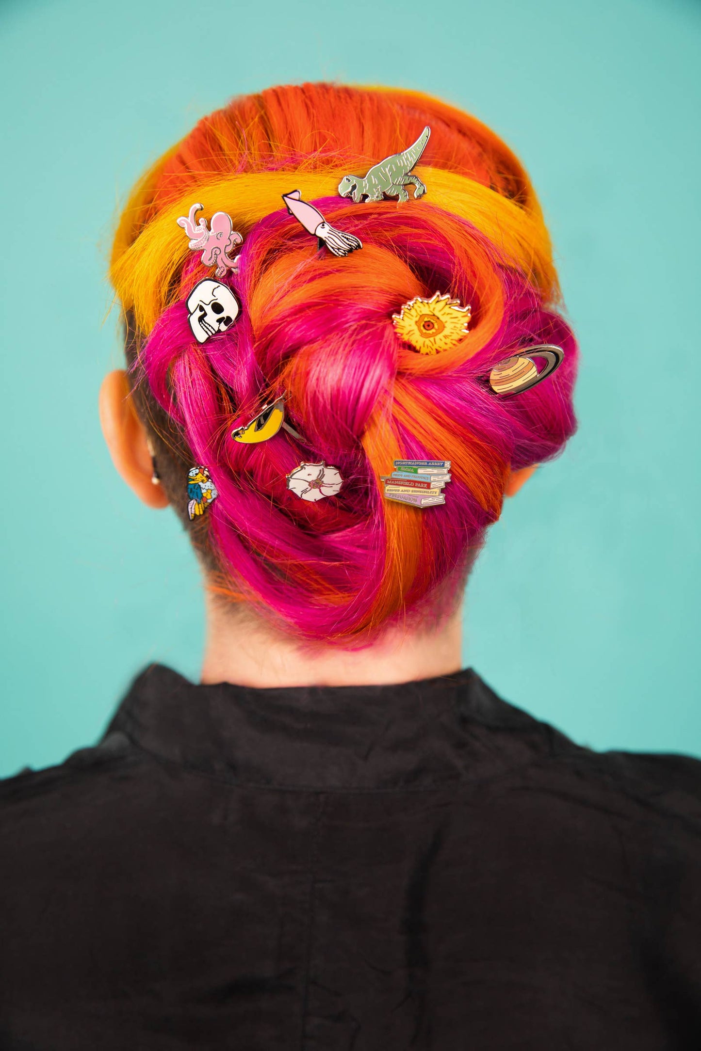 A person with vibrant orange and pink hair in an updo adorned with Dinosaur Hair Pins shaped like a dinosaur, octopus, cat, sun, books, and planets stands against a teal background.