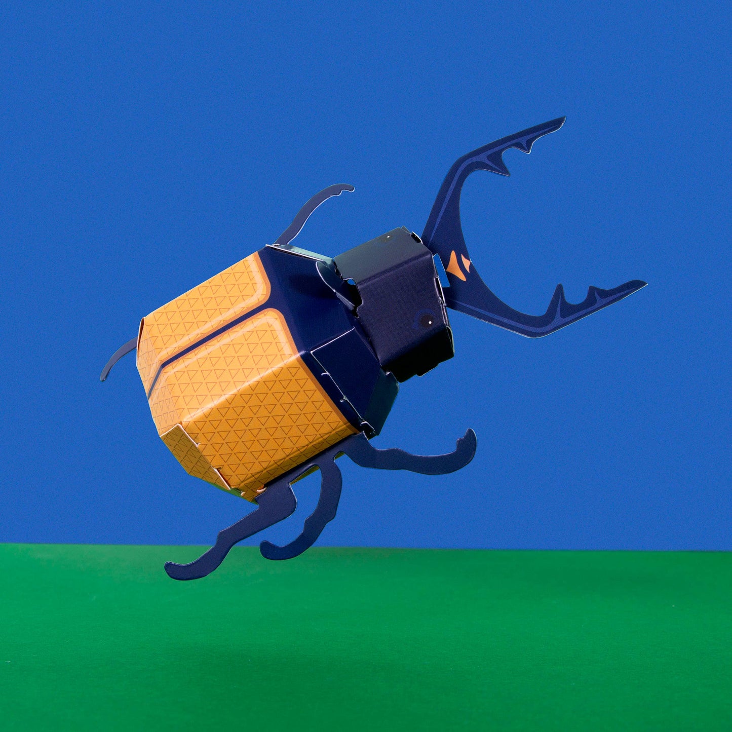 The Create Your Own Super Stag Beetle for Kids features a stylized yellow-orange and blue geometric beetle model displayed on a green surface with a solid blue background.