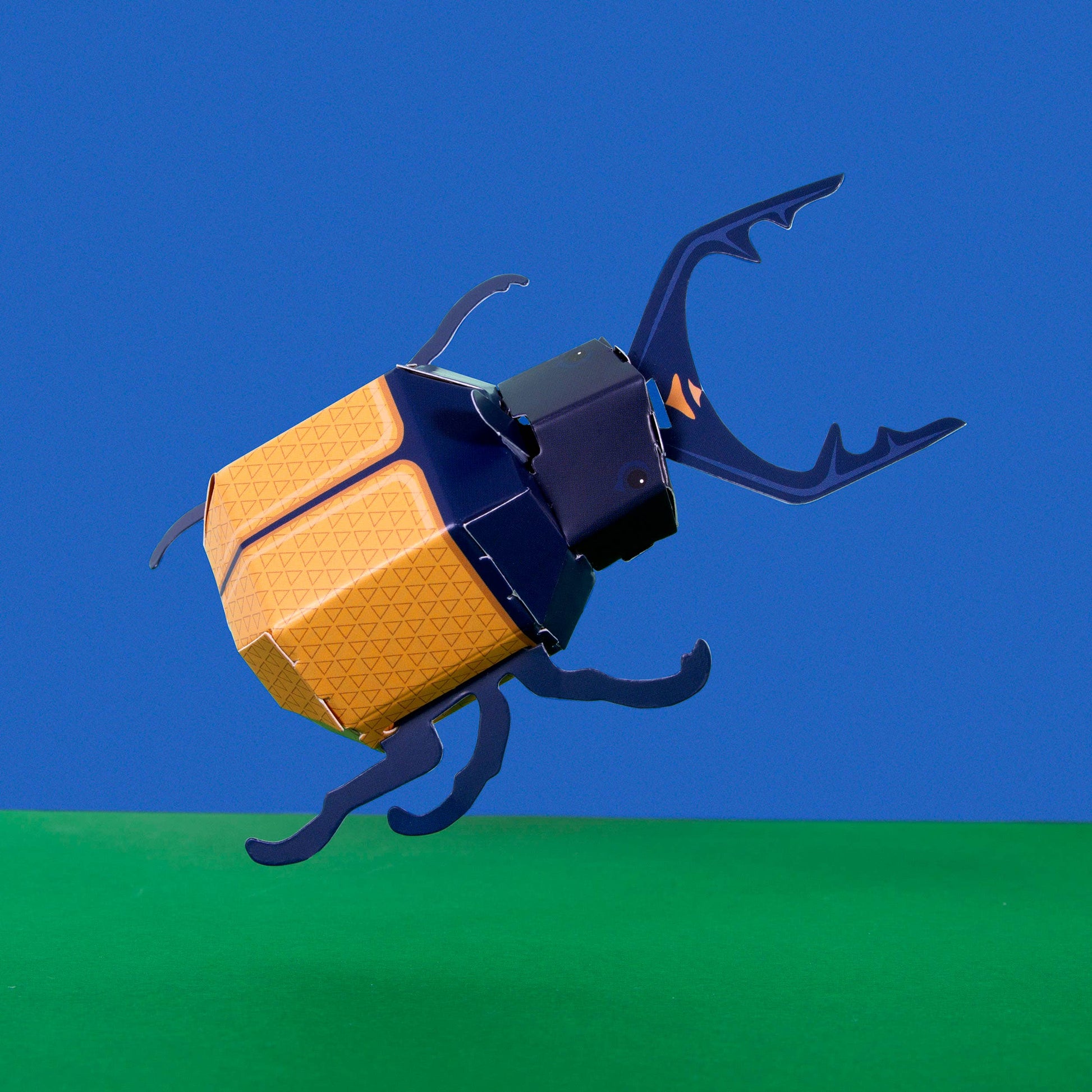 The Create Your Own Super Stag Beetle for Kids features a stylized yellow-orange and blue geometric beetle model displayed on a green surface with a solid blue background.