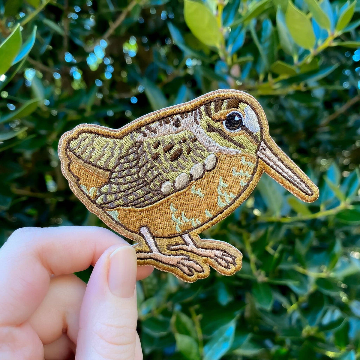 A hand holds an American Woodcock Iron-On Embroidered Patch shaped like a bird with brown and tan plumage and a long beak.