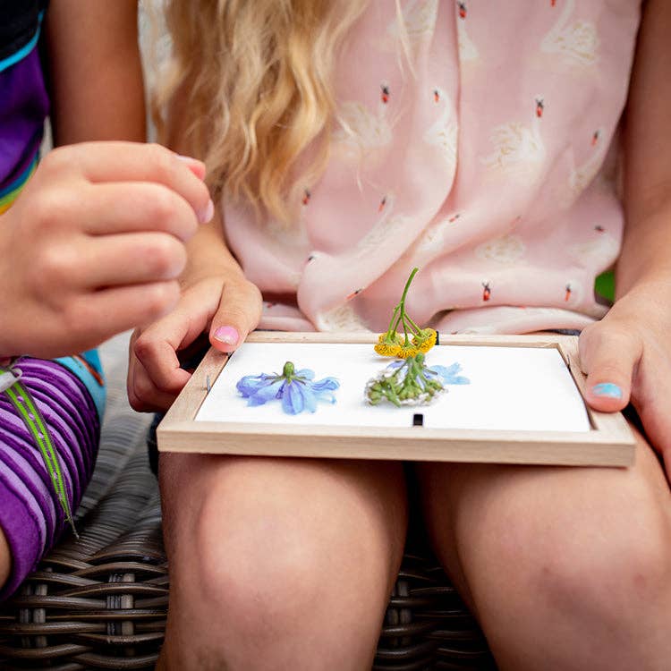 Two children sit close, holding a Huckelberry Pressed Flower Frame with blue and yellow blooms on white blotting paper. One wears a light pink swan-patterned dress and has painted nails.