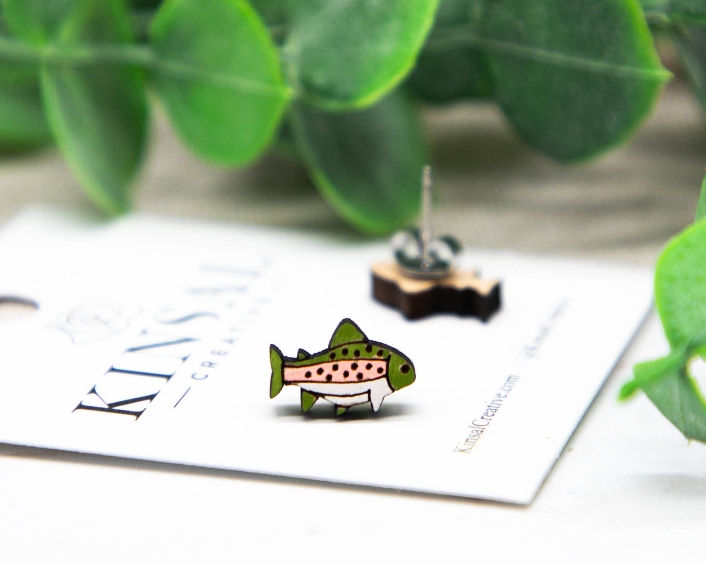 A Trout Fish Wood Stud Earring rests on a white card with green leaves in the background, while the second earring and part of the handmade jewelry branding are also visible.