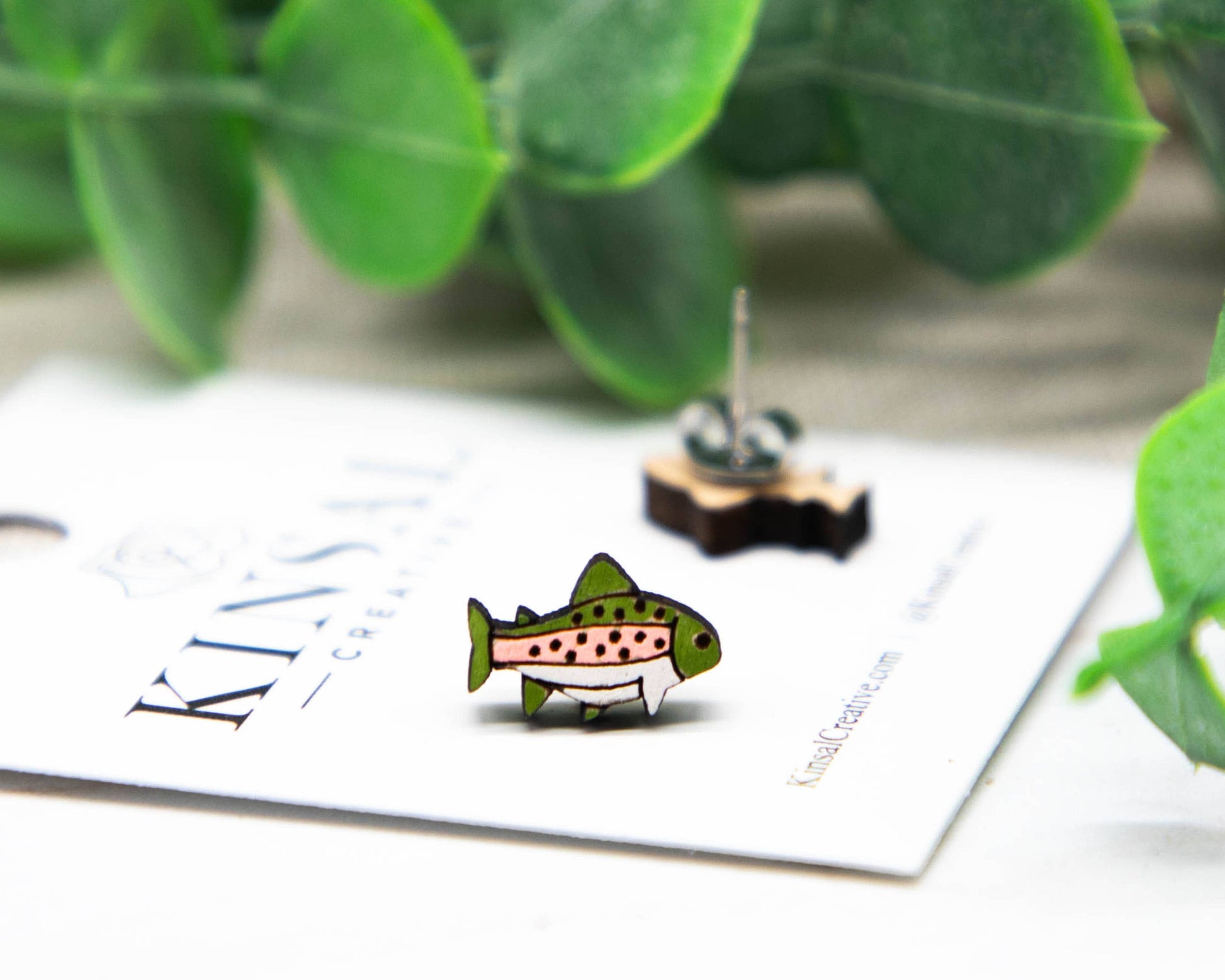 A Trout Fish Wood Stud Earring rests on a white card with green leaves in the background, while the second earring and part of the handmade jewelry branding are also visible.