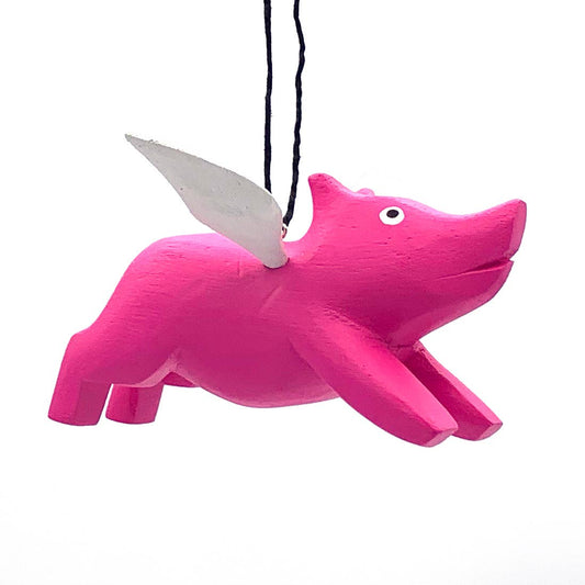 The Flying Pig Balsa Ornament, a hand-carved pink pig with white wings, hangs from a black string and appears to fly against a plain white background.