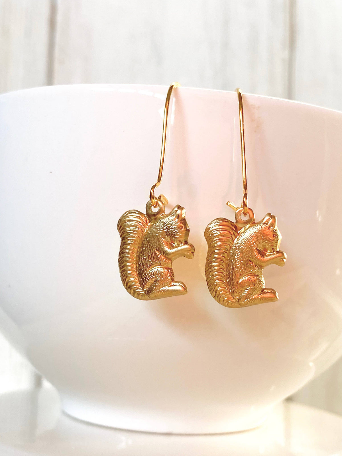 A pair of Woodland Forest Squirrel Earrings, crafted in gold and shaped like adorable squirrels with intricate fur details and bushy tails, hang from hooks. Displayed against a white bowl on a light, neutral background, these charming earrings capture the playful spirit of forest creatures.