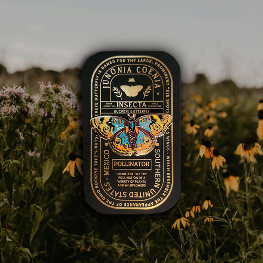 A Buckeye Butterfly Educational Insect Enamel Pin tin, featuring a vibrant butterfly illustration and educational text about pollinators, is nestled among yellow wildflowers in a field, with softly blurred greenery and sky in the background—making it a perfect gift for butterfly enthusiasts.