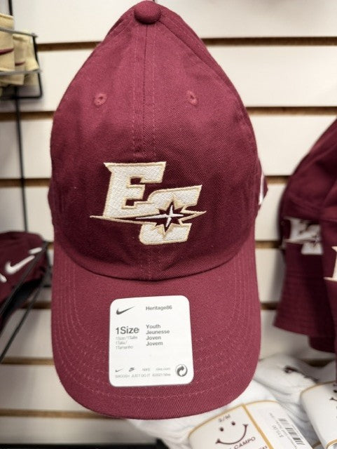 A Nike Youth Cap in maroon, featuring a white embroidered EC logo and star design on the front, is displayed on a rack in a store. A label on the brim identifies it as a youth-sized Heritage86 cap.