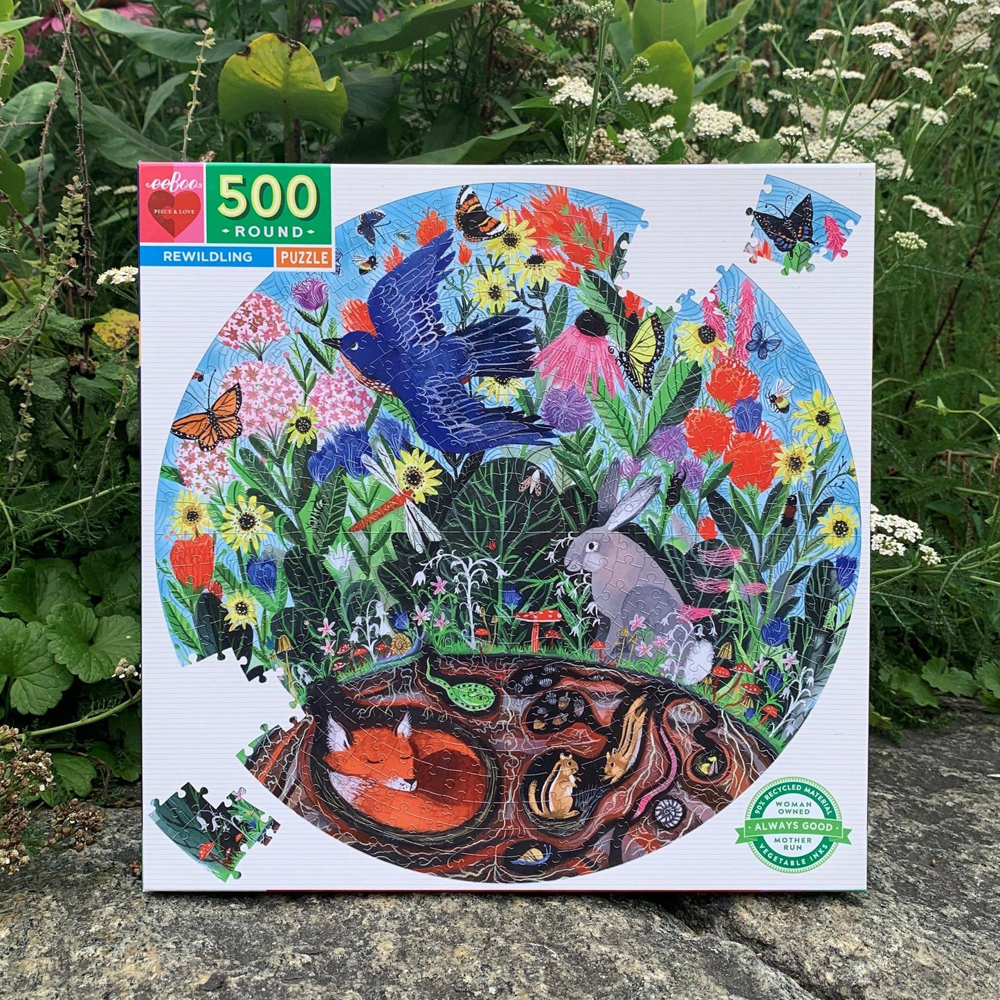 The Rewilding 500 Piece Puzzle is a round sustainable jigsaw featuring a vibrant woodland scene with fox, rabbit, birds, colorful flowers, and butterflies. The box is pictured outdoors on stone amid lush green plants.