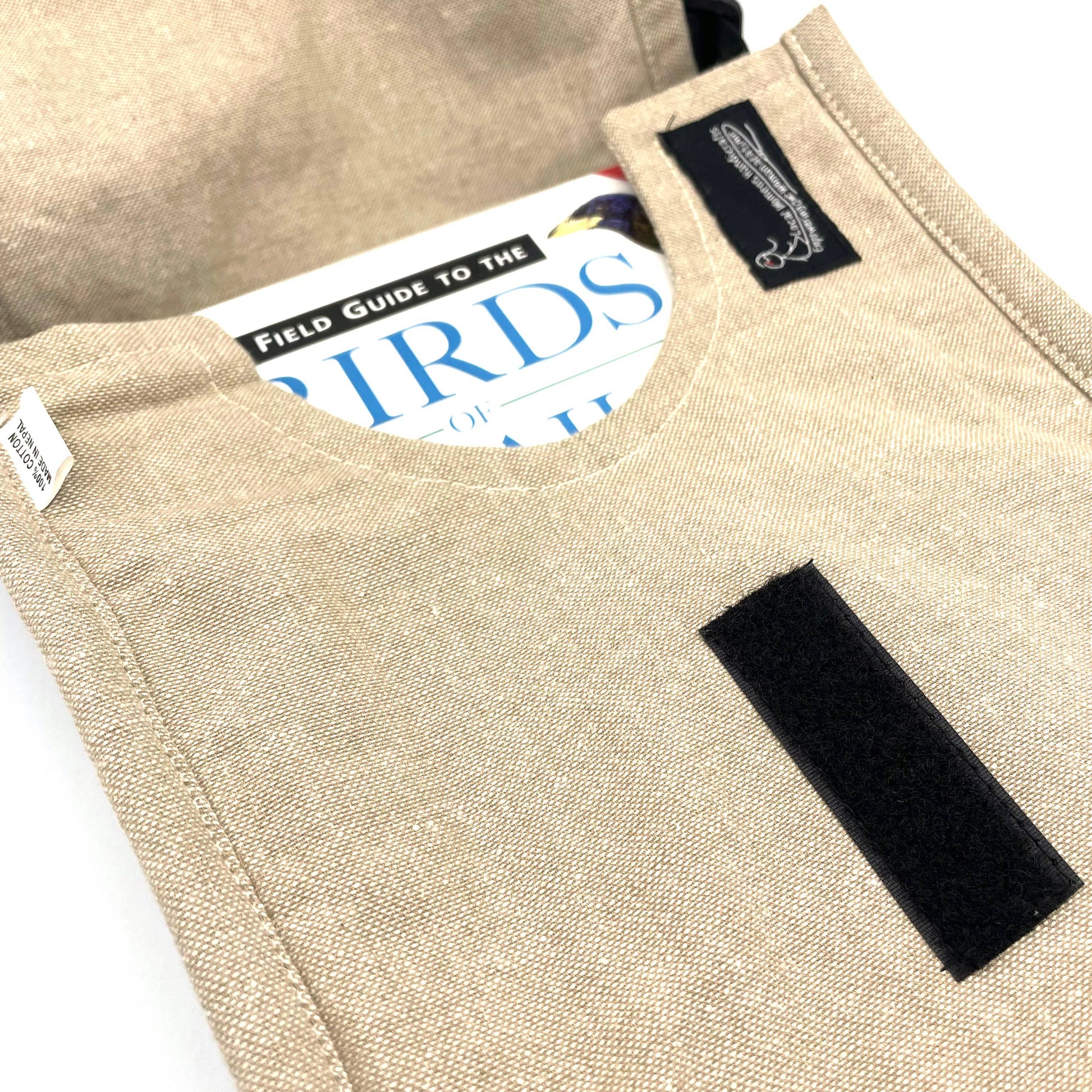A beige linen Luna Moth Field Bag, partially open, reveals a book titled Birds inside. The pouch features a black Velcro strip on the front and a black label sewn inside the flap.